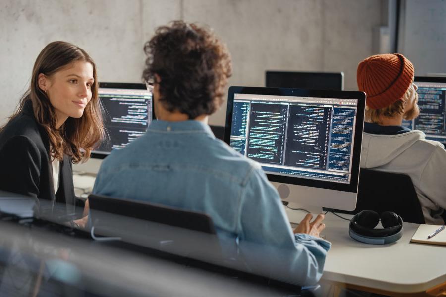 Two CompTIA students sitting in front of a computer and one of the students is talking to a CompTIA Academy instructor.